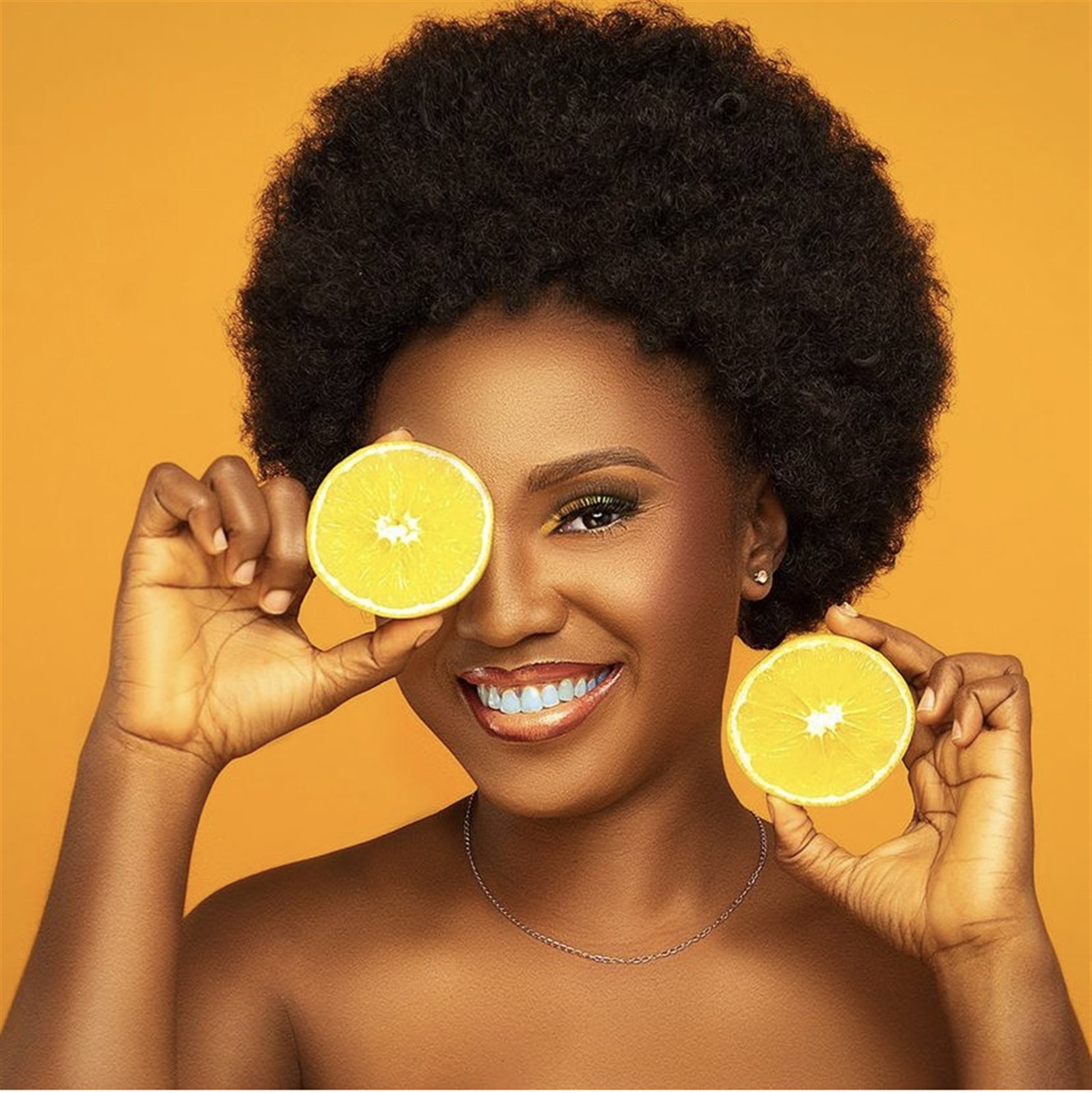 Smiling woman holding orange slices over her eye against an orange backdrop