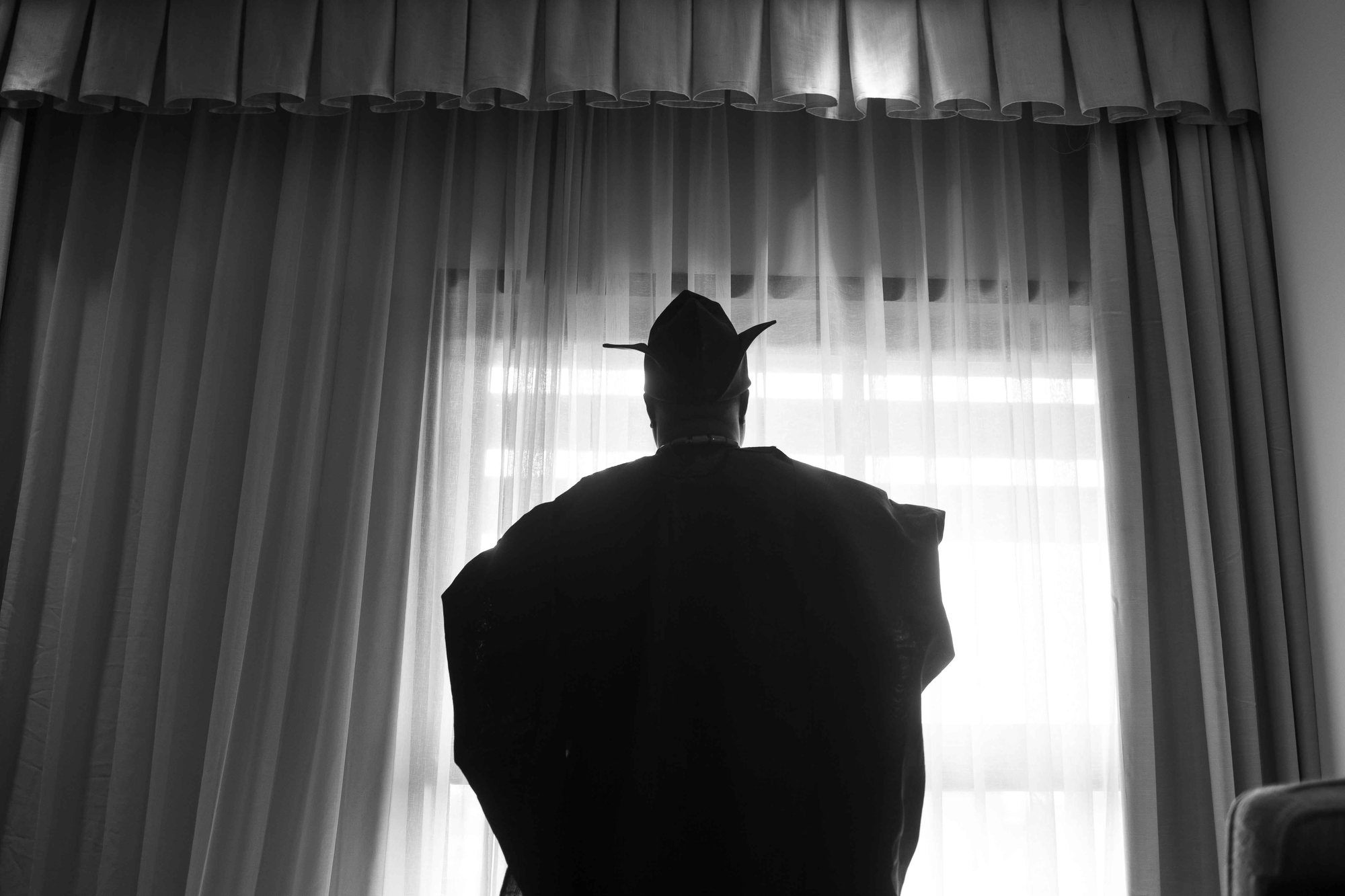 Groom in traditional attire silhouetted against a sheer curtain